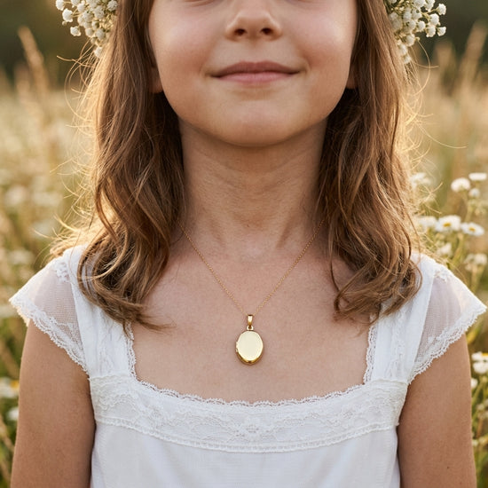 Mädchen trägt goldenes Oval-Medaillon Pure Mini an feiner Kette, weißes Kleid mit Spitze, Blumenwiese im Hintergrund