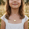 Mädchen trägt goldenes Oval-Medaillon Pure Mini an feiner Kette, weißes Kleid mit Spitze, Blumenwiese im Hintergrund