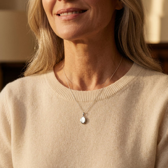 Frau trägt ovales Mini-Medaillon aus Silber an feiner Kette über beigem Pullover, dezent und elegant