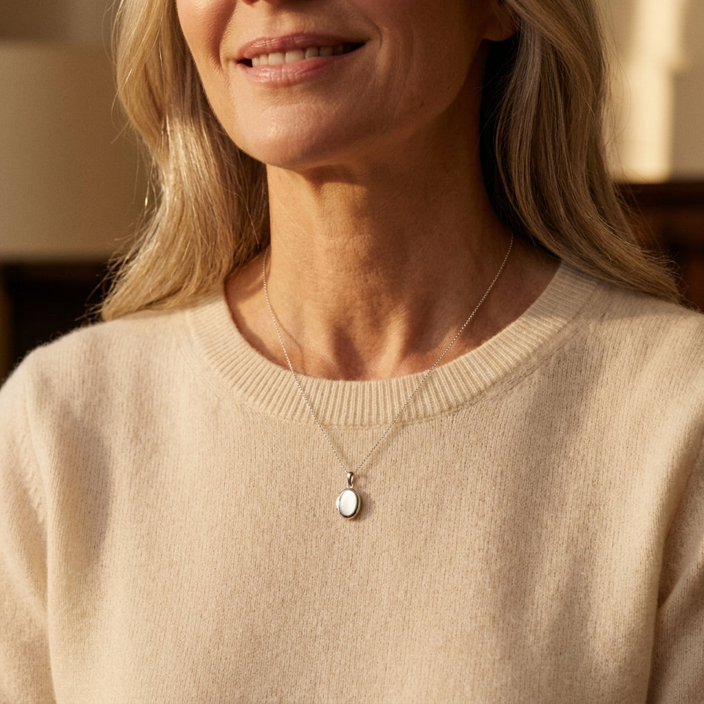 Frau trägt ovales Mini-Medaillon aus Silber an feiner Kette über beigem Pullover, dezent und elegant