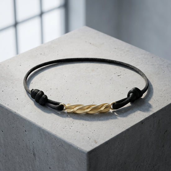 Schwarzes Textilarmband mit gedrehtem Goldelement, liegt auf weißer Steinoberfläche