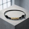 Schwarzes Textilarmband mit gedrehtem Goldelement, liegt auf weißer Steinoberfläche