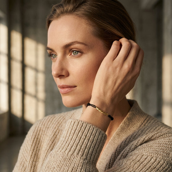 Frau mit blonden Haaren trägt goldenes Armbändchen Breeze mit schwarzen Akzenten am Handgelenk, beiger Strickpullover