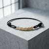 Schwarzes Lederarmband mit goldener gedrehter Spirale als Mittelelement auf grauem Betonwürfel