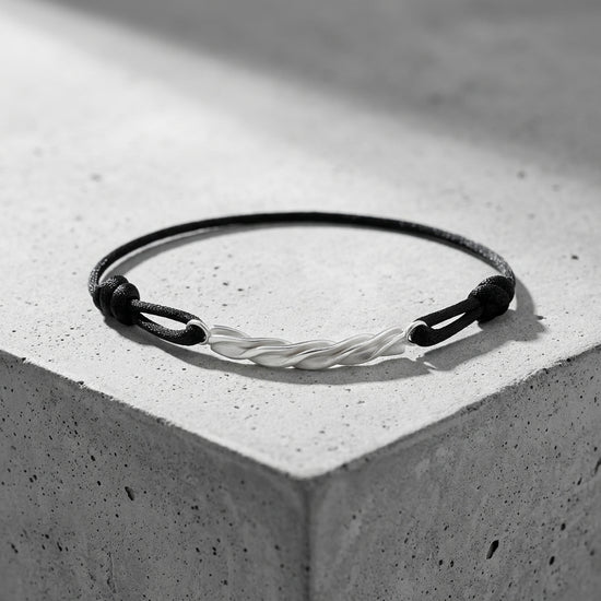 Schwarzes Textilarmband mit gedrehtem Silberelement in der Mitte, auf grauem Betonwürfel präsentiert