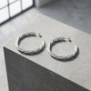 Silberne Creolen Breeze large mit gedrehtem Design auf grauem Betonblock, hochwertige Handarbeit aus Pforzheim