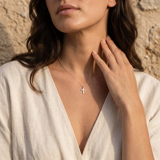 Frau trägt silbernen Kreuz-Anhänger Essence Mini von Aurevia an feiner Kette, minimalistisches Design vor natürlichem Hinterg