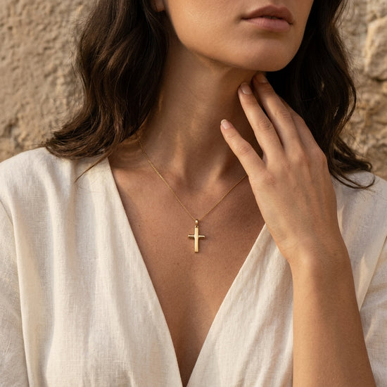 Frau trägt goldenen Kreuz-Anhänger Exelsis an feiner Kette, Hand am Hals, weißes Leinenhemd, natürlicher Look